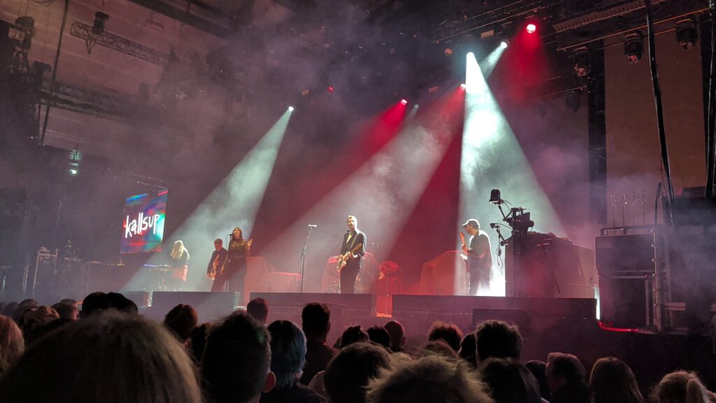 Kallsup on stage at K.B. Hallen in red and white spotlights and smoke, with the band name on the screen